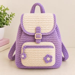 May include: A crochet backpack in lavender and cream yarn. The bag features a cream-colored top flap with a lavender buckle strap, a cream front pocket with a lavender zipper, and a small lavender flower appliqué on the lower right corner.