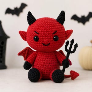 May include: A red crocheted devil doll with black horns, black hands, and black feet. The doll features black plastic eyes, a small smile, red wings, and a pointed tail. It holds a black crocheted trident in one hand, showcasing detailed yarn work.