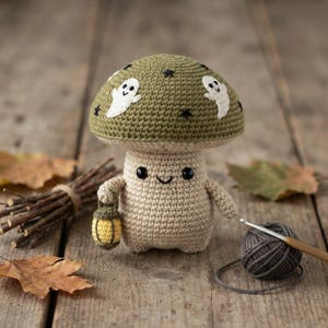 May include: A crocheted mushroom figure with a sage green cap featuring white ghost and black star embroidery. The beige body has black bead eyes, a stitched smile, and holds a small yellow lantern. The figure is crafted with yarn.