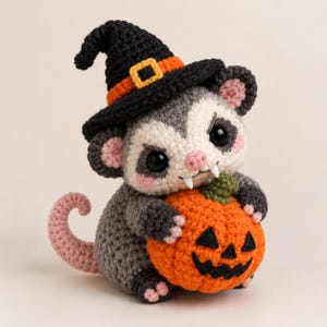 May include: A crocheted stuffed animal of a grey and white opossum wearing a black witch hat with an orange band and buckle. The opossum has black eyes, pink ears, and two white fangs, and it is holding a small orange crocheted jack-o'-lantern.