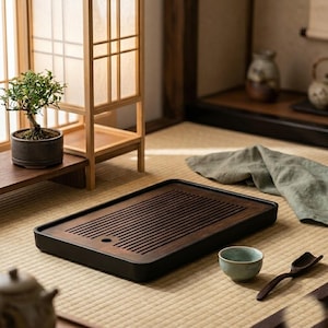 Puede incluir: Una bandeja de té rectangular de madera oscura con una superficie de listones y un orificio de drenaje circular. La bandeja tiene un marco oscuro y liso, situada sobre una esterilla tejida junto a una pequeña taza de cerámica azul claro y una cuchara.