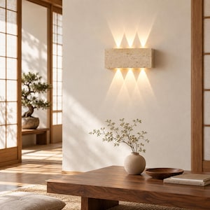 May include: A rectangular wall sconce made of beige travertine stone with a porous texture. The fixture is mounted on a plain wall and emits warm, bright light in three distinct beams directed both upward and downward from the top and bottom edges.