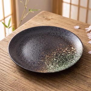 以下が含まれることがあります： 黒い質感のある表面を持つ円形の陶磁器の皿。表面は金と緑の斑点模様へとグラデーションで変化しています。皿にはわずかに盛り上がった縁があり、全体的にマットな仕上げで、まだら模様の外観が特徴です。
