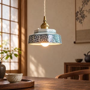 May include: A pendant light featuring a hexagonal ceramic shade with a white base and blue geometric patterns. The fixture includes a brass-toned metal socket and a twisted fabric cord, with a single light bulb glowing from the center.