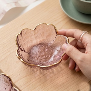 Può includere: Una piccola ciotola in vetro a forma di fiore con una tonalità rosa traslucida e una superficie testurizzata e ondulata. Il bordo della ciotola è rifinito con un sottile profilo color oro metallizzato, che mette in risalto le curve organiche.