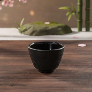 Könnte beinhalten: Eine kleine, schwarze Teetasse aus Gusseisen mit einem strukturierten, erhabenen Diagonalmuster auf der Außenseite. Die Tasse hat einen glatten, abgerundeten Rand, ein tiefes, dunkles Inneres und einen konischen Boden, der auf einer Holzoberfläche steht.