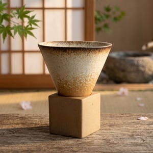 May include: A ceramic coffee dripper with a conical shape featuring a speckled cream and brown gradient glaze. The dripper rests securely on a solid, matte tan square base, showcasing a textured finish and a wide, open top rim for brewing.