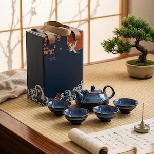 May include: A ceramic tea set featuring a dark blue and black gradient glaze, including a teapot with a lid and four matching small bowls. The set is presented with a tall, rectangular blue gift box decorated with a crane and wave illustration.
