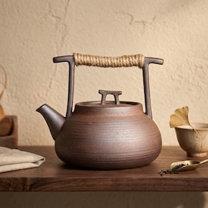 Op de afbeelding: Een donkerbruine keramische theepot met een ronde vorm, een korte tuit en een plat deksel met een klein handvat. De theepot heeft een hoog, gebogen metalen handvat dat is omwikkeld met natuurlijk jutekoord voor een goede grip.