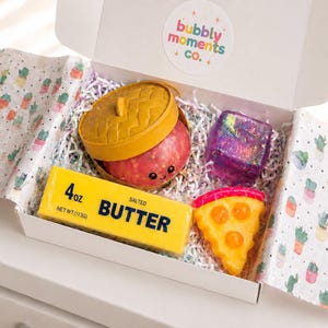 May include: A white gift box containing four items: a yellow rectangular bar labeled 4oz salted butter, a round yellow lidded container, a purple iridescent cube, and a yellow pizza slice. The box features a bubbly moments co. logo sticker.
