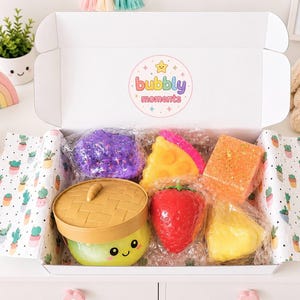 May include: A white cardboard box containing various squishy toys, including a purple textured sphere, a yellow pizza slice, a red strawberry, a square orange block, a yellow triangular shape, and a green container with a woven lid and face.