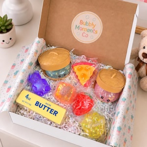 May include: A cardboard gift box containing various sensory toys, including two round containers with woven lids, a yellow butter-shaped block labeled 4oz, a purple bear, a pizza slice, a red strawberry, and a yellow sphere, all nestled in shredded paper.