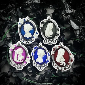 May include: Five oval-shaped decorative pins featuring white silhouette profiles of characters set against vibrant backgrounds of blue, black, magenta, and red. Each pin is framed in silver with ornate floral accents and small colored beads.