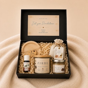 Könnte beinhalten: Eine schwarze Geschenkbox mit einem Holzuntersetzer mit Herzanhänger, einer kleinen Flasche ätherischem Öl, einer ZIRBE-Kerze, einem kleinen Stoffbeutel mit Anhänger und einem Glas Badesalz Zirbe, inklusive einer Karte mit Zeit zum Durchatmen.