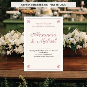 May include: A rectangular wedding invitation card with a cream background and a thin pink border featuring small floral corner accents. The text reads: Alexandra & Michael, Saturday, the Twelfth of June, Two Thousand Twenty-Seven, Four O'Clock.