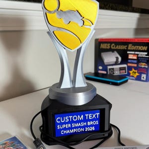 May include: A 3D-printed trophy featuring a silver base and a silver and yellow shield-shaped topper. The black rectangular base displays a blue screen with the white text: CUSTOM TEXT, SUPER SMASH BROS, CHAMPION 2026. A black power cord is attached.