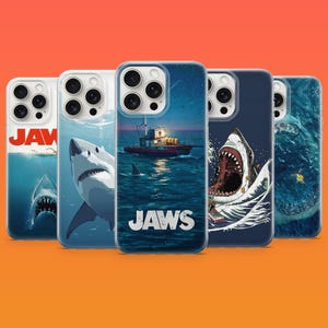 May include: Five smartphone cases featuring Jaws movie imagery. Designs include a shark approaching a swimmer, a shark head, a boat at sea with the text JAWS, a shark with an open mouth attacking a boat, and an aerial view of a shark in the ocean.
