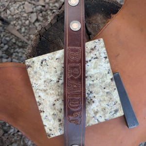 May include: A dark brown leather strap featuring the name BRANDT embossed vertically in a serif font. The strap is secured with four circular metal rivets, two at the top and two at the bottom, and includes decorative stitching along the edges.