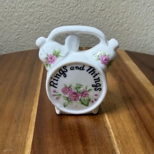 May include: A white ceramic trinket holder shaped like a vintage alarm clock with a handle at the top. The front features pink rose floral patterns and the black text Rings and Things printed in a circular arc across the center of the piece.
