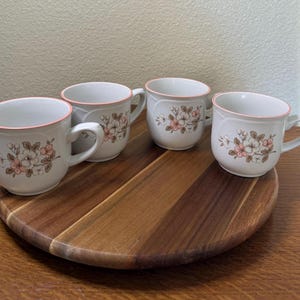 May include: Four white ceramic mugs with a delicate floral pattern featuring pink and brown blossoms. Each mug has a thin pink rim and a curved handle. The floral design is centered on the front of each mug, showcasing detailed petals and leaves.