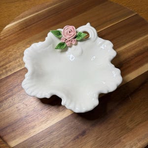 May include: A white ceramic trinket dish with a ruffled edge and a small, three-dimensional pink rose accent with green leaves attached to the rim. The dish has a glossy finish and a small circular hole near the floral decoration on the top edge.
