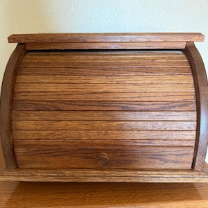May include: A medium brown wooden bread box featuring a roll-top tambour door design. The storage container is constructed from horizontal wooden slats and includes a small, circular wooden knob centered on the bottom slat for opening the door.