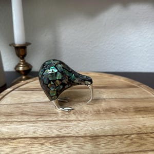 May include: A kiwi bird figurine featuring a body covered in iridescent abalone shell mosaic tiles with a dark base. The bird has a long, curved silver-toned beak and matching silver-toned feet, standing in a profile position on a flat surface.