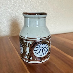 May include: A small ceramic vase with a speckled off-white glaze and a wide neck. The lower body features a decorative band with brown circular motifs containing dark blue floral patterns. The base is cylindrical with a smooth, finished rim.