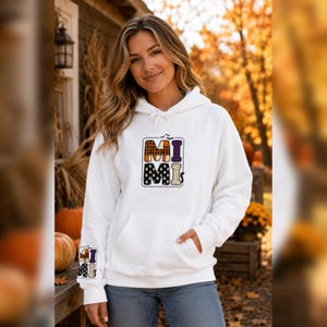Peut inclure: Un sweat-shirt à capuche blanc avec un motif sur la poitrine et la manche gauche formant le mot MIMI en lettres capitales. Les lettres sont remplies de motifs à carreaux orange et noirs, violets et noirs avec de petits fantômes.