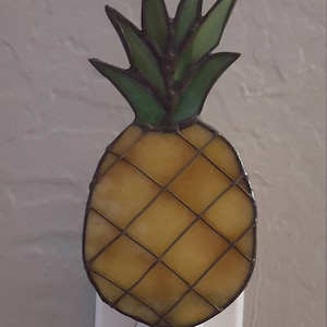 May include: A stained glass pineapple night light plugged into an electrical outlet. The pineapple is yellow with a green top and brown accents.