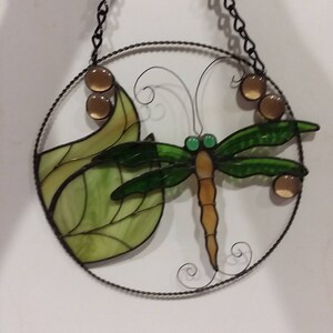 May include: A stained glass dragonfly with green wings and a brown body is surrounded by a leaf and brown glass beads. The dragonfly is hanging from a metal chain.