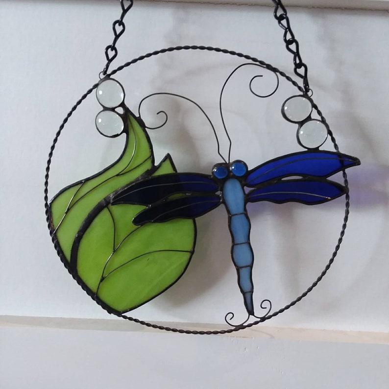 May include: A stained glass dragonfly with blue wings and a green leaf, hanging from a black chain in a circular frame.