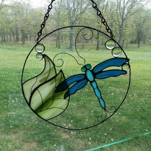 May include: A stained glass dragonfly suncatcher featuring a blue and green dragonfly design and a green leaf. The suncatcher is suspended from a chain and has clear glass beads.