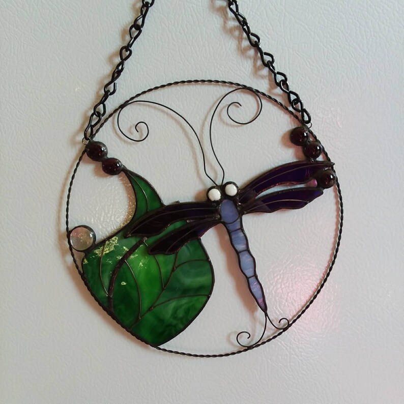 May include: A stained glass dragonfly with purple wings and a green leaf, hanging from a black chain in a circular frame.