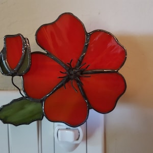 May include: A stained glass night light shaped like a red poppy flower with a green leaf and a bud. The night light is plugged into a standard electrical outlet.