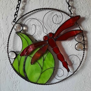 May include: A stained glass dragonfly with red wings and a green leaf, hanging from a black chain.