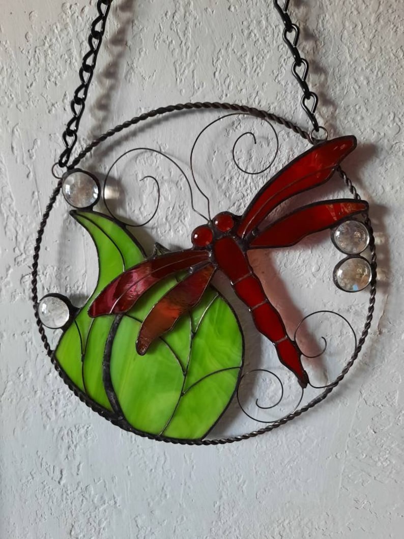May include: A stained glass dragonfly with red wings and a green leaf, hanging from a black chain.