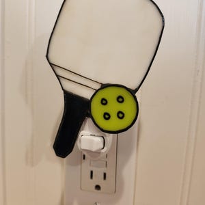 May include: A stained glass night light shaped like a pickleball paddle with a green ball. The paddle is white with a black handle and trim. The ball is green with four black dots. The night light plugs into a standard wall outlet.