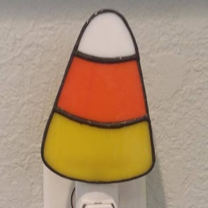 May include: A stained glass candy corn night light plugged into an electrical outlet. The night light is yellow, orange, and white.