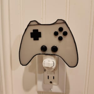 May include: A white and black stained glass night light shaped like a video game controller. The controller has a black outline, a black plus sign, and four black buttons. It is plugged into a white electrical outlet.