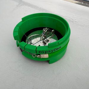 May include: A bright green circular fishing accessory with the text DipsyDen printed on the side. The device features a hollow center containing a metallic silver fishing lure with hooks and a swivel mechanism attached to a thin fishing line.