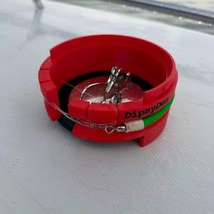May include: A red circular fishing line winder with the text DipsyDen printed on the side. The device features a bright green accent band and contains a silver metal fishing lure with a clear monofilament line wrapped around the interior.