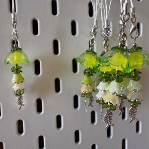Puede incluir: Una colección de colgantes para teléfono con cuentas translúcidas en forma de flor verde y amarilla dispuestas en niveles. Cada colgante tiene un cierre de mosquetón plateado y termina con un pequeño dije plateado en forma de espina de pescado.