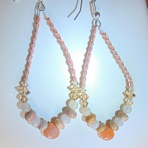 May include: A pair of teardrop-shaped earrings featuring a woven peach-colored cord frame. The lower section of each earring is adorned with a sequence of faceted clear crystals, smooth pale beads, and textured tan and orange stone beads.