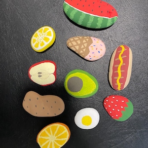 Op de afbeelding: Een verzameling van tien gladde rivierstenen, met de hand beschilderd met diverse voedselontwerpen, waaronder een watermeloenplak, citroenplak, ijshoorntje, appelplak, halve avocado, hotdog, aardappel, aardbei, sinaasappelplak en een gebakken ei.