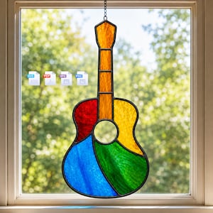 May include: A stained glass suncatcher shaped like an acoustic guitar, featuring a multi-colored design with amber, red, yellow, blue, and green glass panels held together by black metal cames, hanging in front of a window with four file icons.