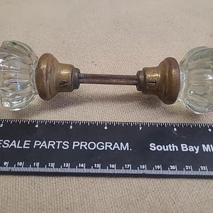 May include: A vintage door knob set featuring two clear, fluted glass knobs attached to brass-toned metal bases and a central connecting spindle. A black ruler is positioned underneath, showing the assembly measures approximately 5.5 inches long.