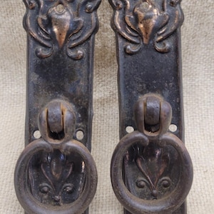 May include: Two rectangular metal drawer pulls with a dark, weathered finish and copper-toned highlights. Each pull features an ornate, raised floral design at the top and a circular ring handle attached to a central mount, with visible screw holes.