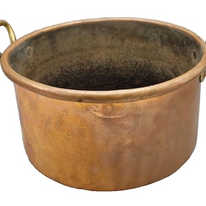 May include: A round copper pot with a weathered, darkened patina on the exterior and interior surfaces. The vessel features two curved brass handles securely attached to opposite sides of the rim with visible rivets, creating a functional design.