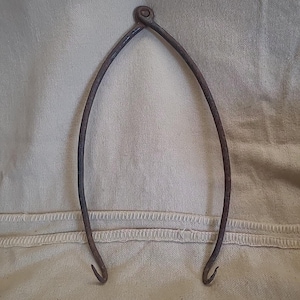 May include: A dark brown, weathered metal caliper tool featuring two curved arms joined at a central pivot point. The arms taper down to sharp, inward-curving points at the bottom, showing signs of oxidation and age on the textured metal surface.